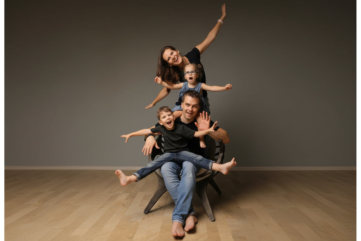 A family of four posing dynamically on a dark chair against a plain gray wall. The mother is sitting on the father's shoulders with her arms raised, while the young boy sits on the father's lap and the toddler stands on the mother's lap, all smiling and waving.